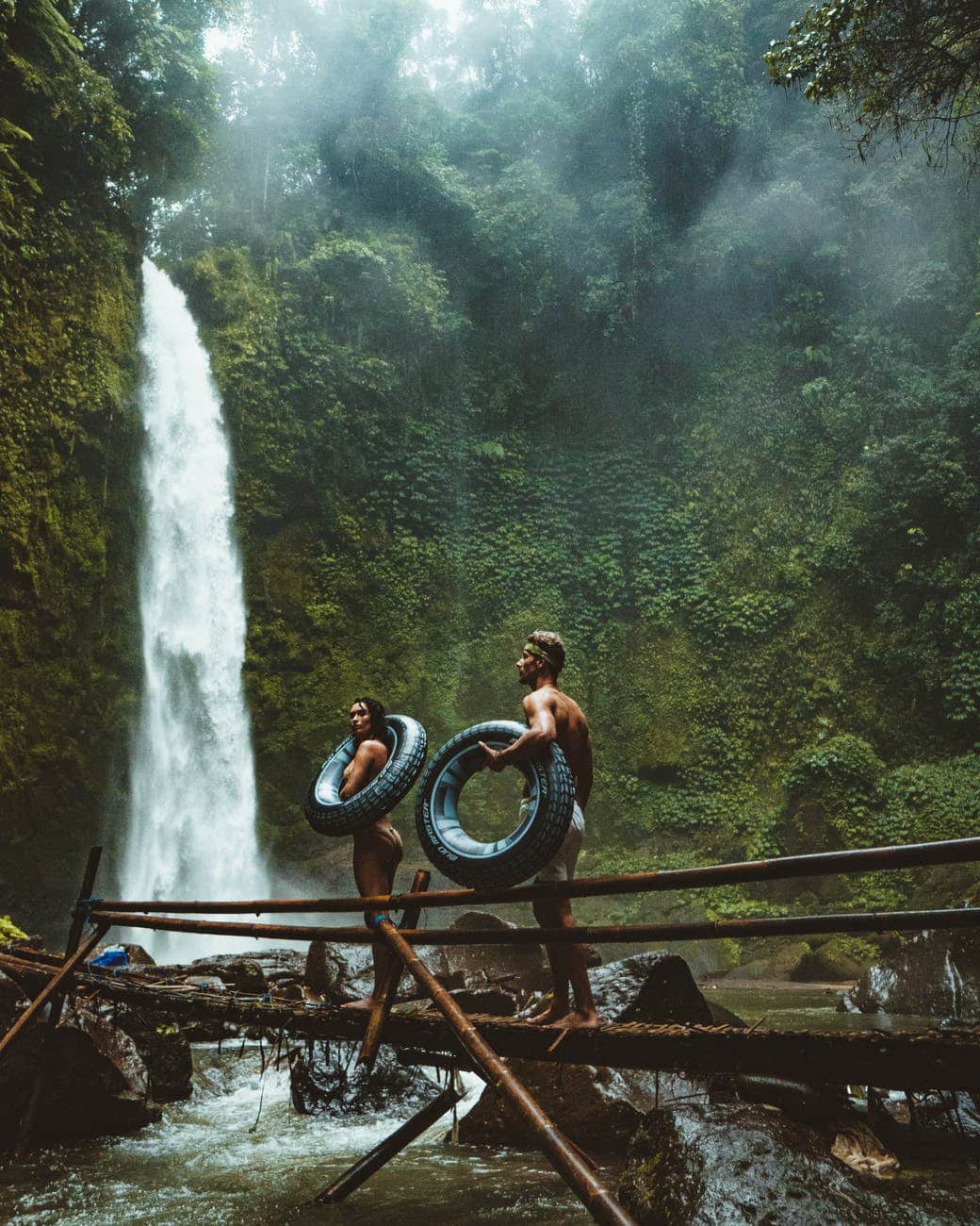 two person carrying black inflatable pool float on brown wooden bridge near waterfalls