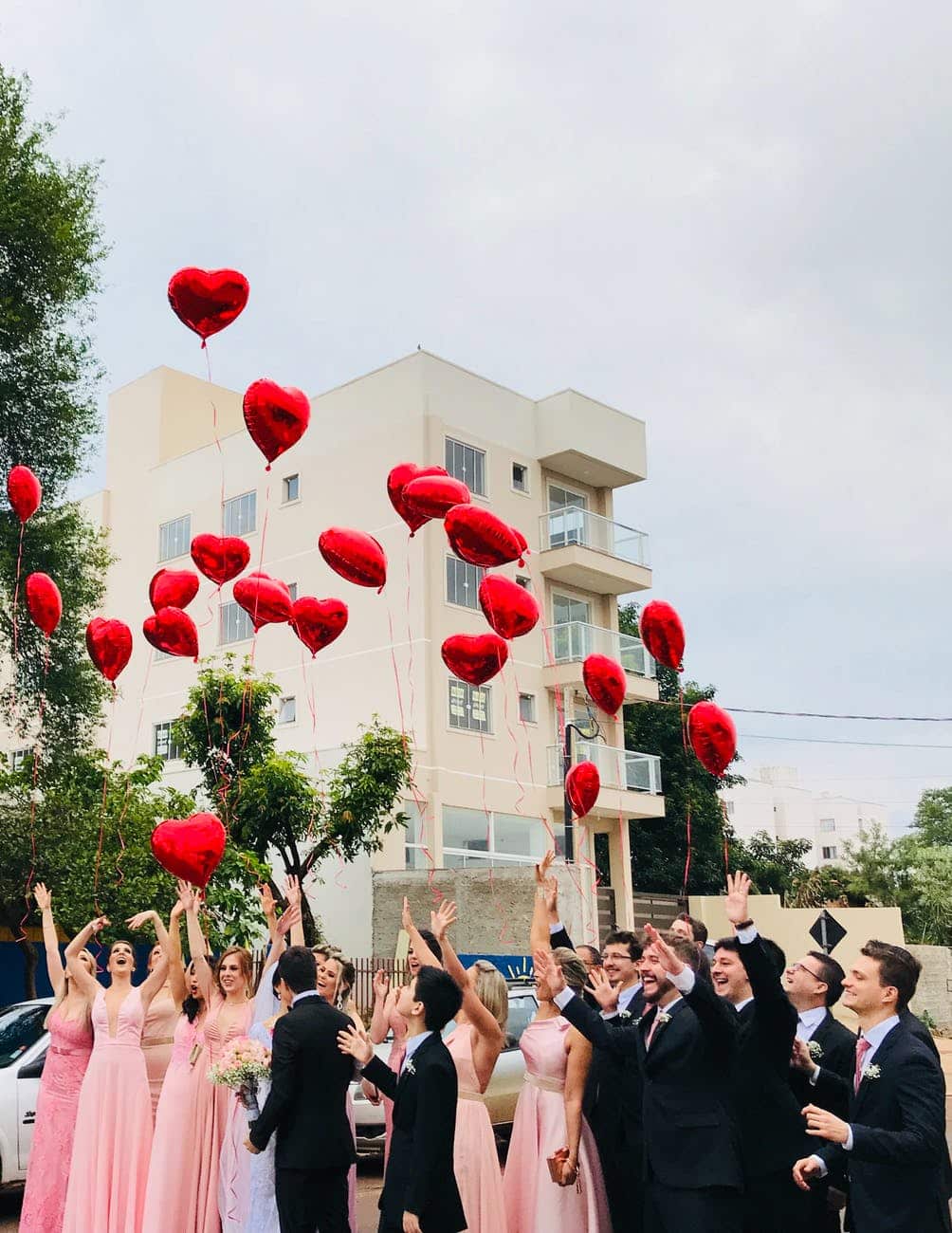 women wearing pink dresses and men wearing black suit jacket and pants raising hands with red heart balloons