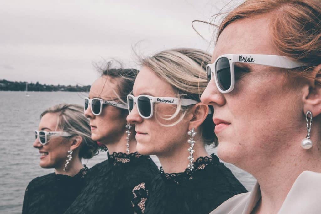 stylish bride and bridesmaids in sunglasses standing on sea embankment on wedding day