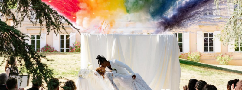 photo de la fin de la cérémonie du mariage de Melvine Malard avec Justine où les mariées s'embrassent avec en fond un feu de jour coloré
